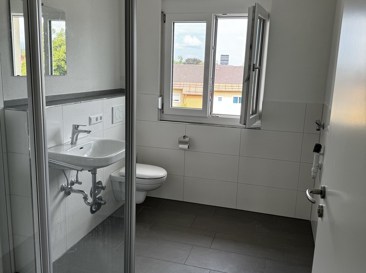 Modernes Bad mit WC, Dusche, schwarzem Fliesenboden und Fenster mit Sichtschutzglas.